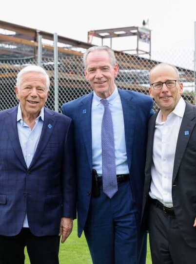 Three men in business attire stand smiling on grass in front of a construction site with a metal fence and steel framework visible in the background.