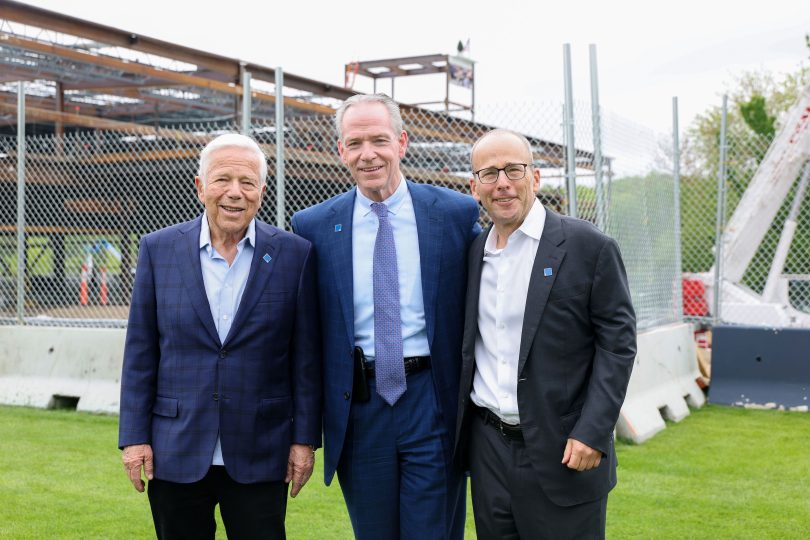 Three men in business attire stand smiling on grass in front of a construction site with a metal fence and steel framework visible in the background.