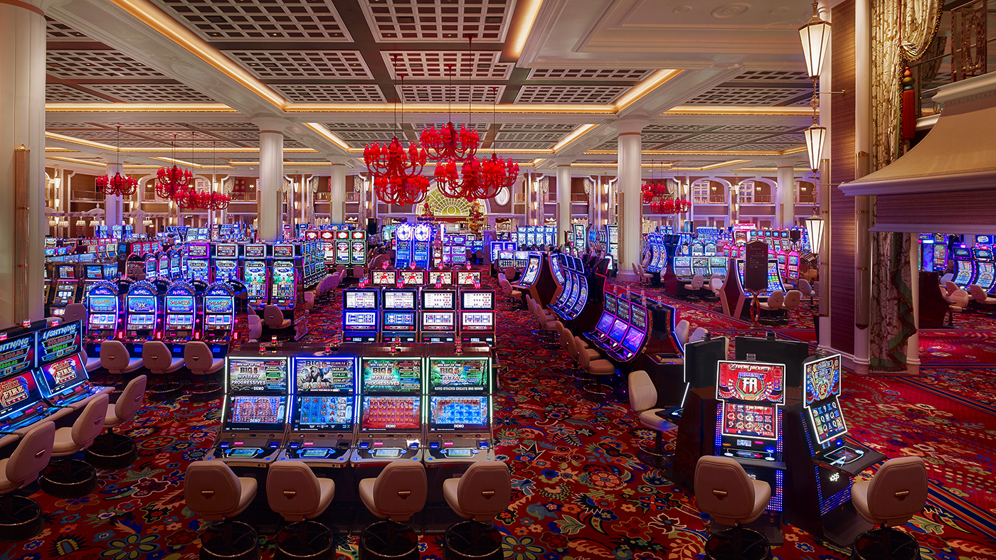 A brightly lit casino floor filled with rows of colorful slot machines, red chandeliers hanging from the ceiling, and patterned carpet throughout the space. No people are visible.