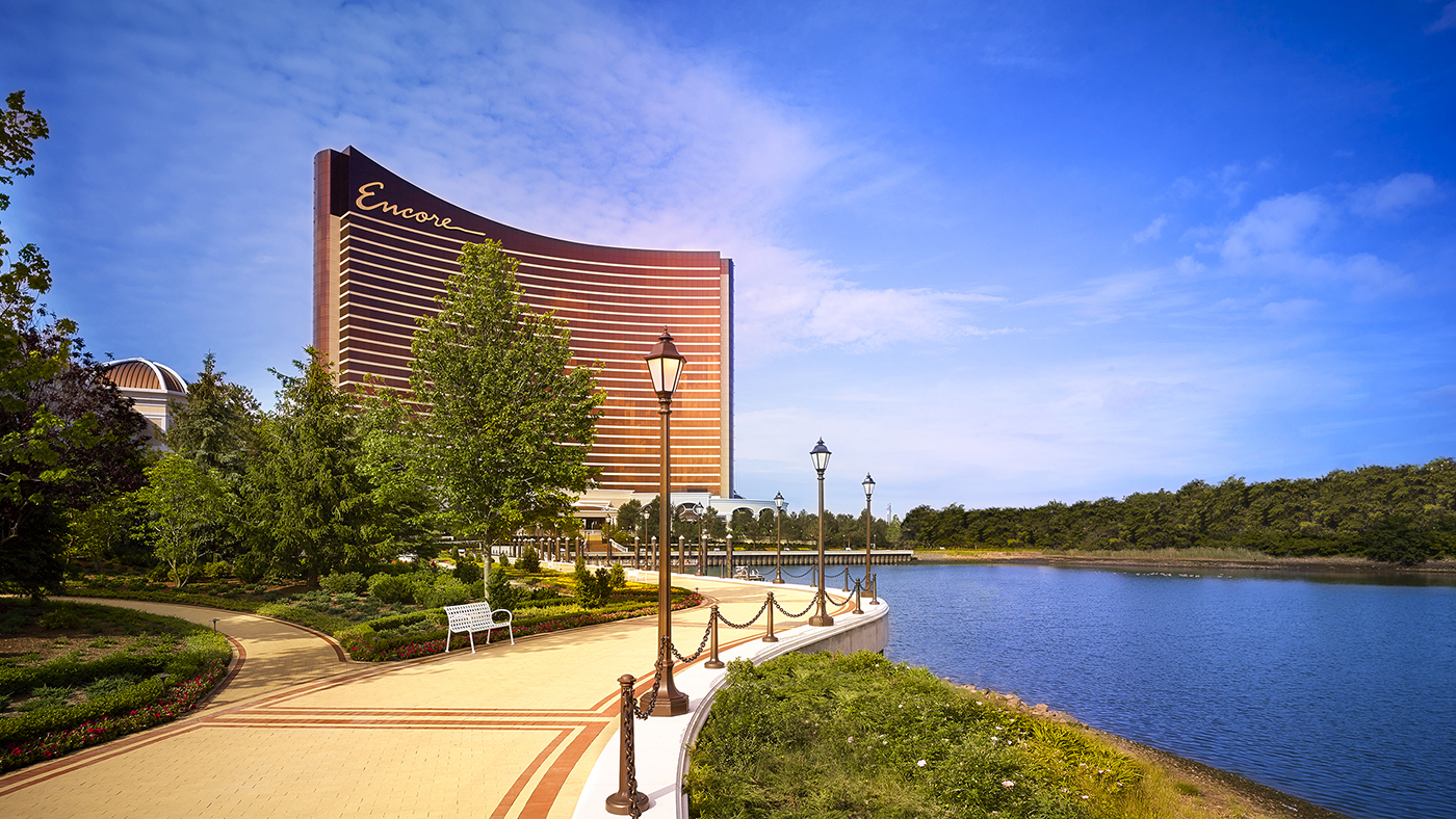 A tall, curved hotel building labeled Encore stands by a landscaped path, green trees, street lamps, benches, and a calm body of water under a bright blue sky.