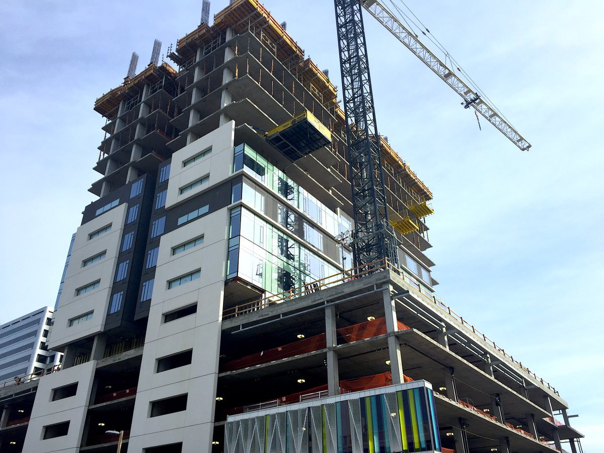 A multi-story building under construction, with exposed concrete floors and scaffolding. A tall crane stands beside the structure, and construction materials are visible on several levels. The sky is clear and blue.