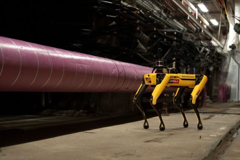 A yellow Boston Dynamics robot dog walks along an industrial corridor next to a large pink pipe, surrounded by cables and equipment.