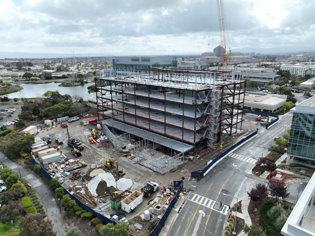 Aerial view of a multi-story steel building under construction, surrounded by construction vehicles and materials, with nearby office buildings, roads, and a body of water in the background.