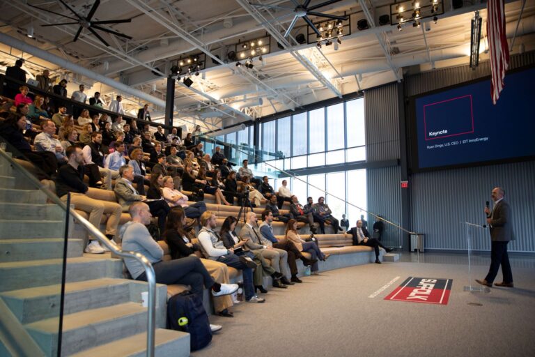 A speaker addresses a large audience seated in stadium-style bleachers inside a modern, well-lit auditorium. A presentation slide titled Keynote is displayed on a large screen behind the speaker.
