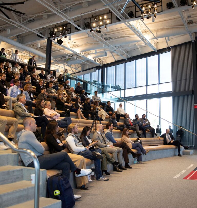 A speaker addresses a large audience seated in stadium-style bleachers inside a modern, well-lit auditorium. A presentation slide titled Keynote is displayed on a large screen behind the speaker.