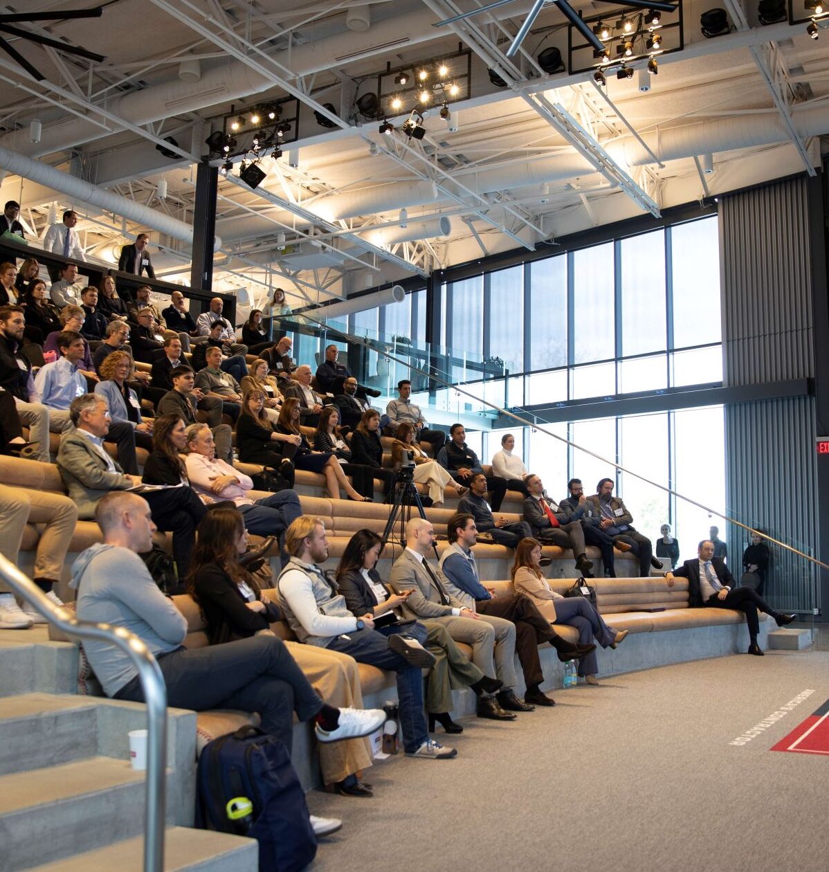 A speaker addresses a large audience seated in stadium-style bleachers inside a modern, well-lit auditorium. A presentation slide titled Keynote is displayed on a large screen behind the speaker.