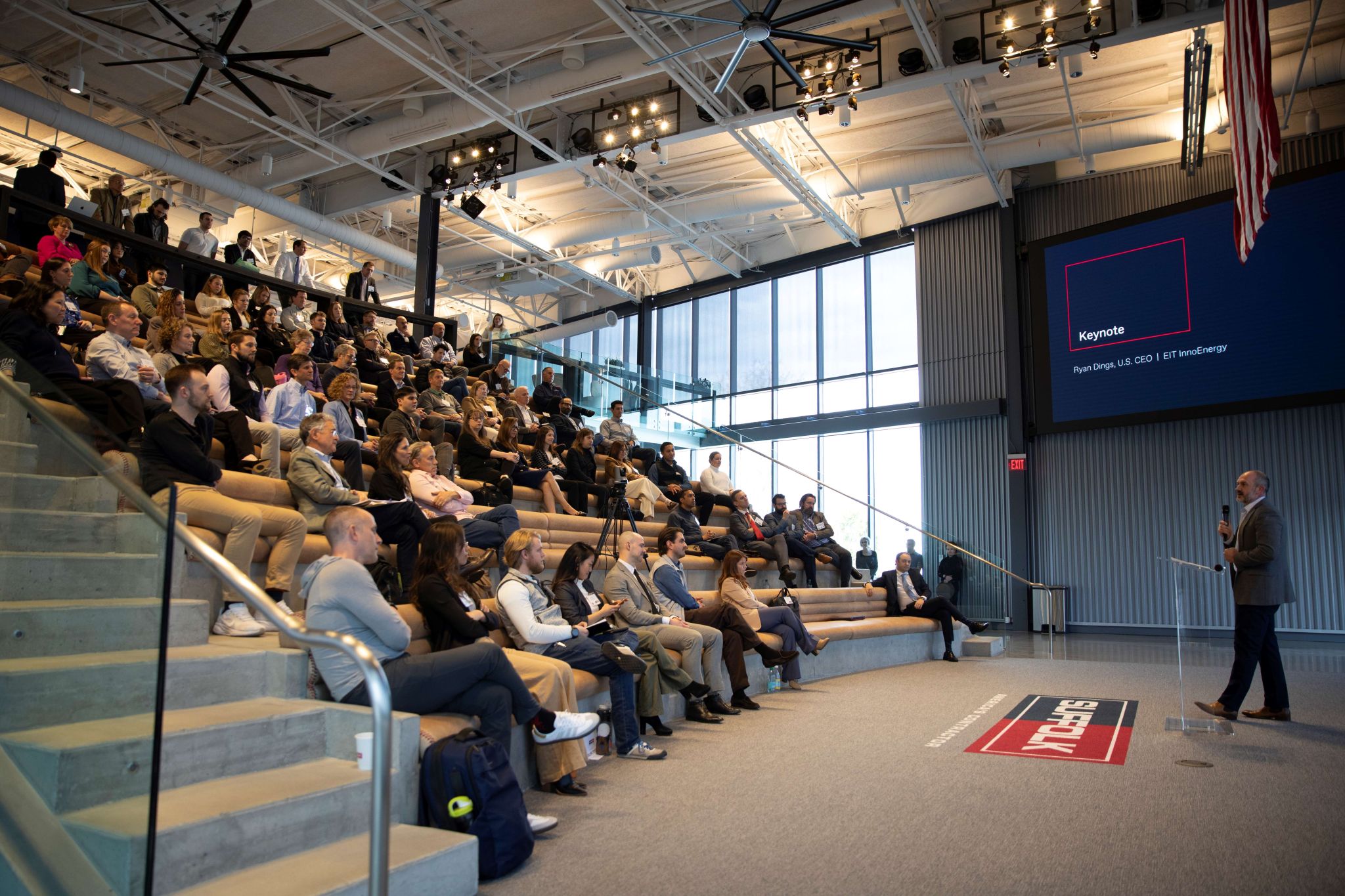 A speaker addresses a large audience seated in stadium-style bleachers inside a modern, well-lit auditorium. A presentation slide titled Keynote is displayed on a large screen behind the speaker.