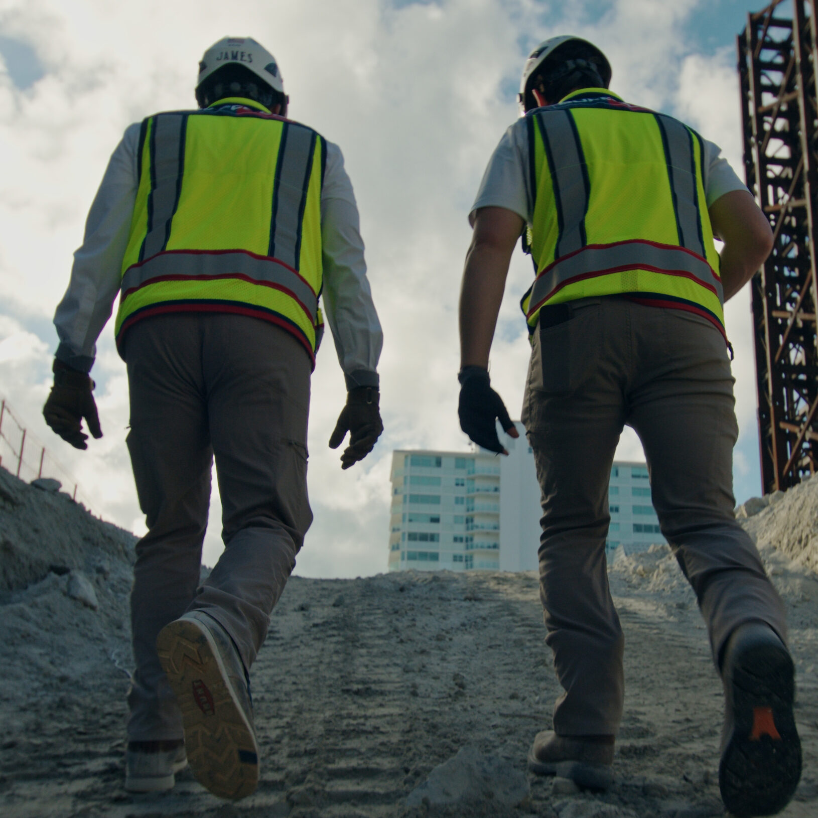Two construction workers in safety vests and helmets walk up a dirt slope at a construction site, highlighting the diverse careers in construction, with a tall building structure and cityscape visible in the background under a cloudy sky.