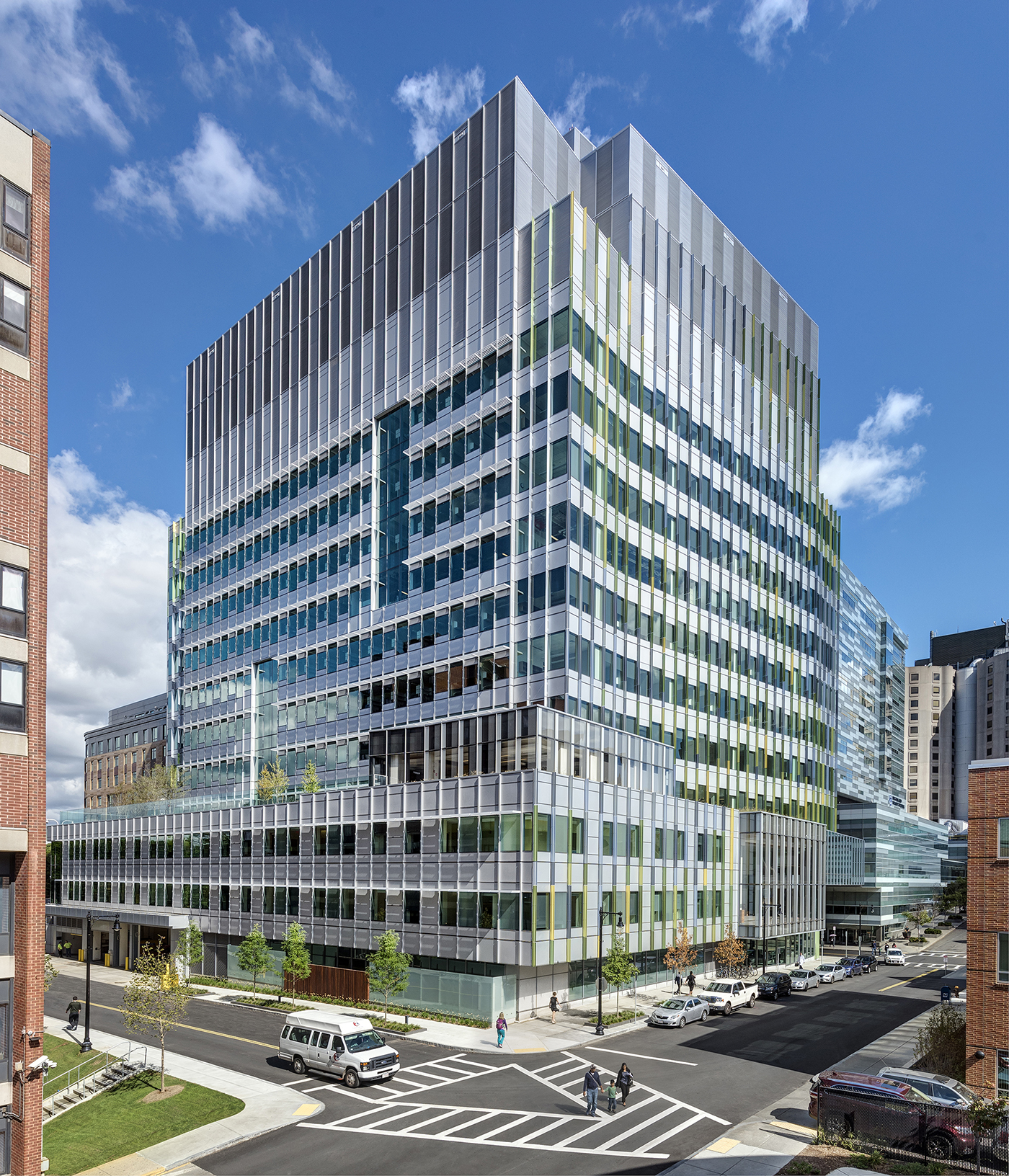 A modern glass office building stands at a busy city intersection with people walking on sidewalks and cars, including a white van, driving on the street under a blue sky with scattered clouds.