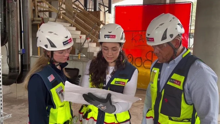 Three construction workers wearing safety helmets and vests examine blueprints together at a building site, with scaffolding and construction materials visible in the background.