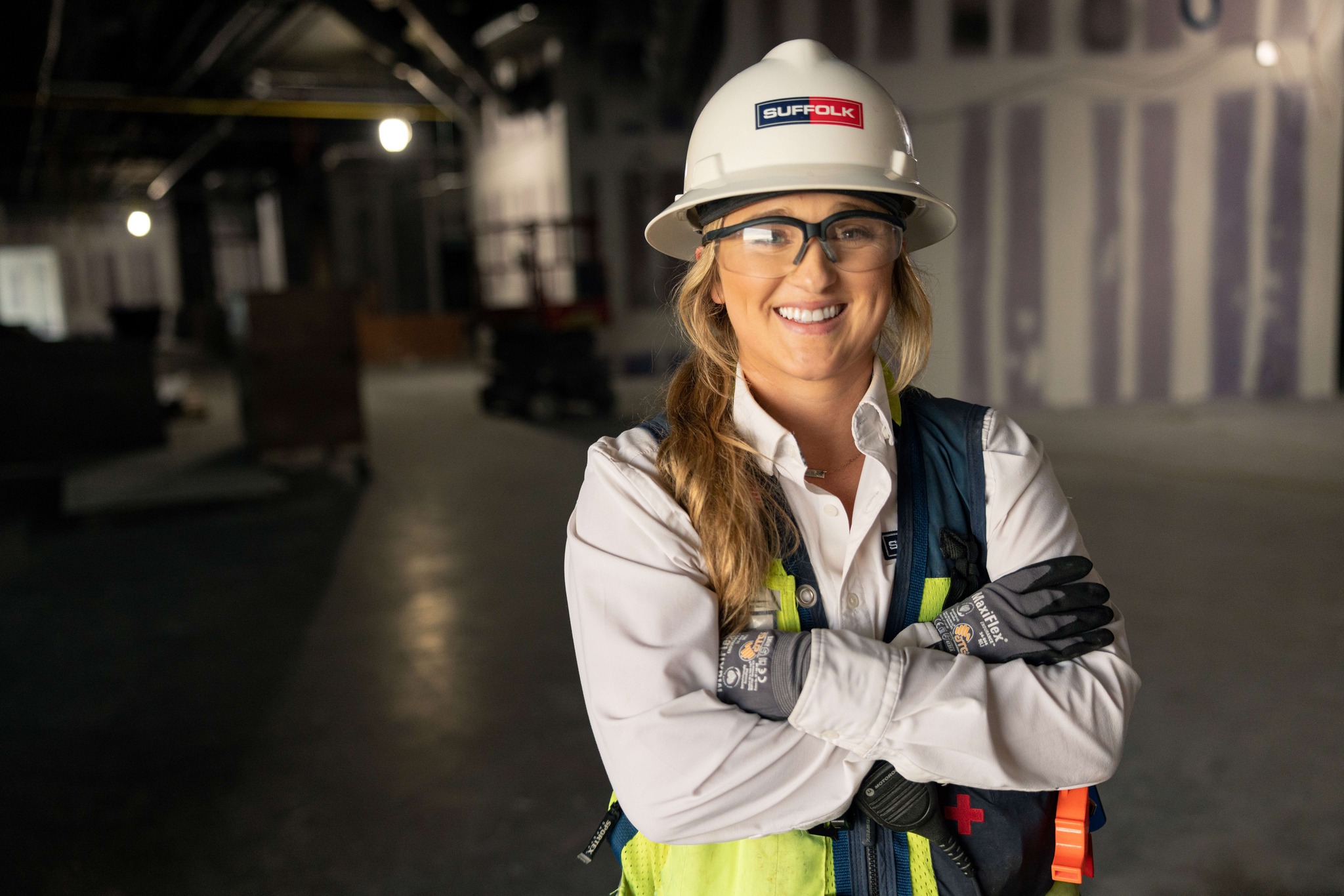 A smiling construction worker wearing a white hard hat, safety glasses, gloves, and a reflective vest stands with arms crossed inside a building under construction.