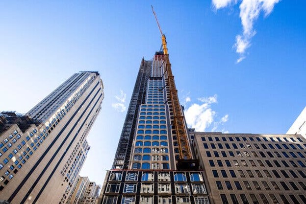 A tall skyscraper under construction, surrounded by other high-rise buildings, with a large yellow crane extending upwards against a bright blue sky with scattered clouds.