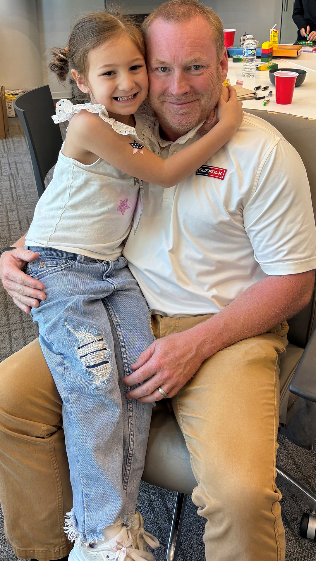 A young girl hugs a smiling man sitting on a chair, both enjoying the benefits of quality time together. The girl wears a white top and ripped jeans, while the man sports a white shirt and khaki pants in a bright indoor setting.