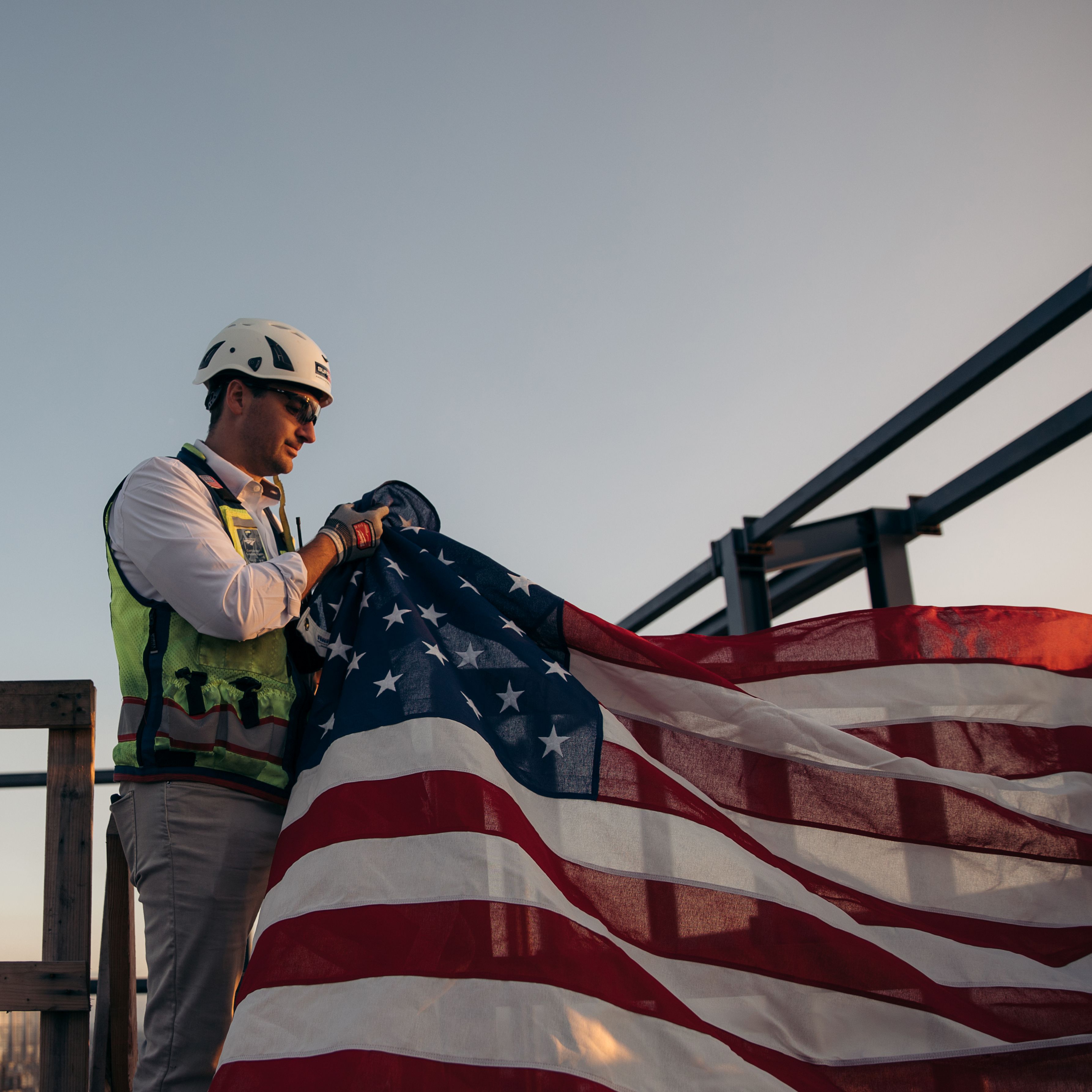 suffolk worker in safety gear holding an american flag during sunset