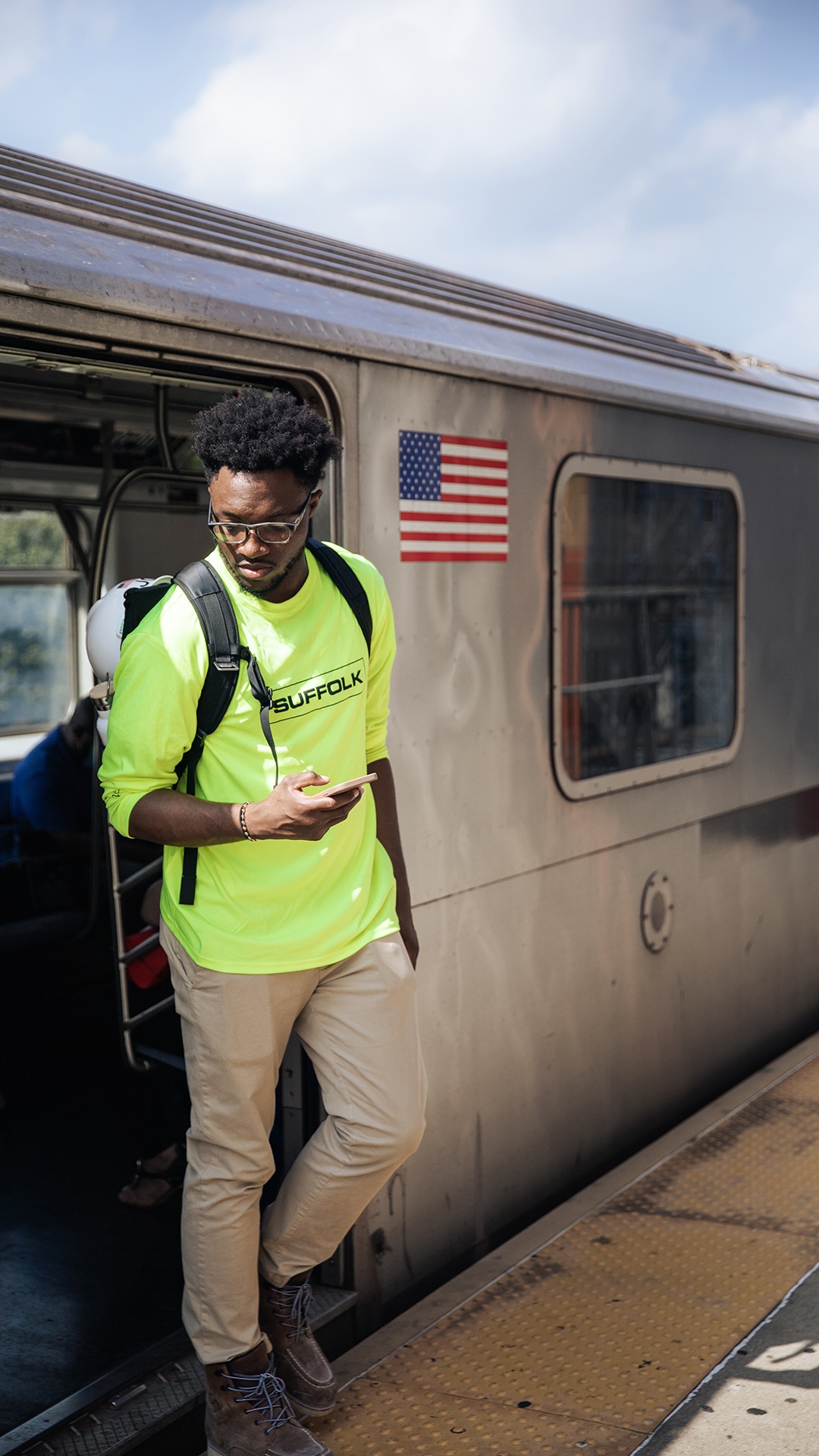 A man in a neon yellow “Suffolk” shirt stands at the open door of a subway train, enjoying the benefits of staying connected on his phone. He has a backpack and sunglasses, and the American flag is visible on the train.
