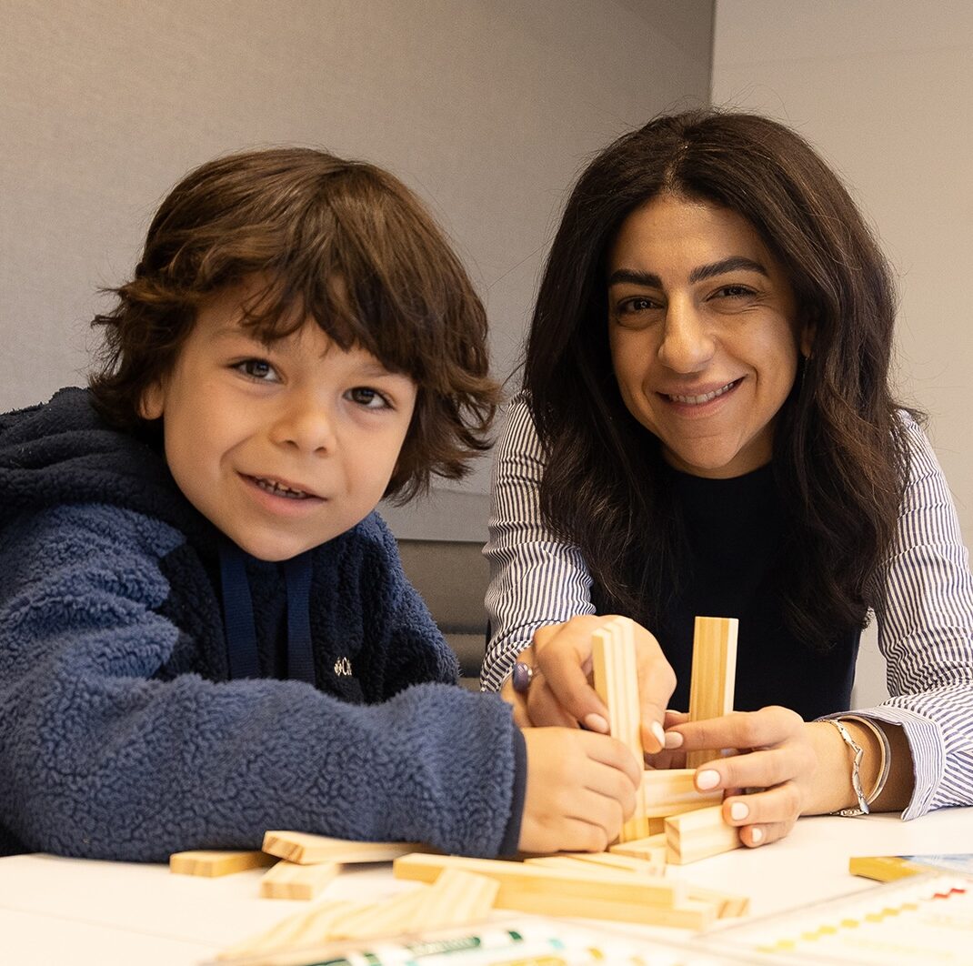 A smiling woman and a young boy enjoy the benefits of creative play as they build with wooden blocks at a table, colorful markers scattered in the foreground. Both are looking at the camera.