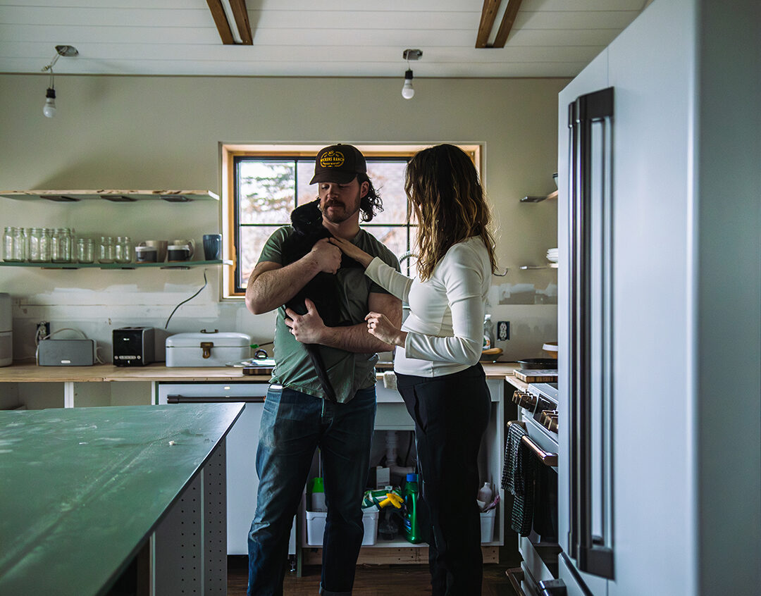 A man holding a black cat stands barefoot in a modern kitchen next to a woman in black pants and boots. They face each other, enjoying the benefits of cozy home living, with shelves and appliances visible in the background.