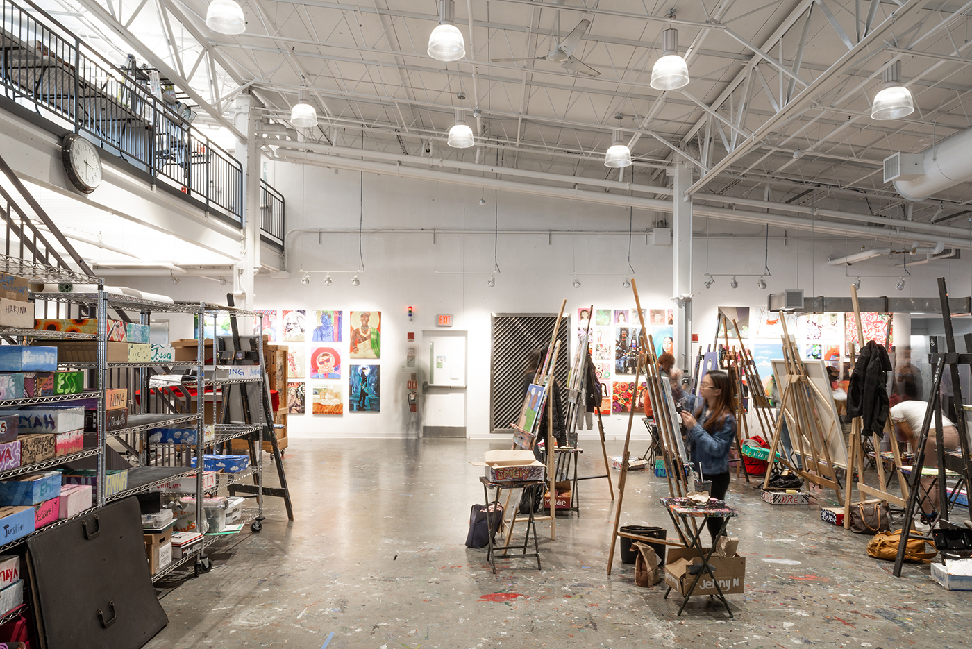 A spacious, brightly lit art studio with high ceilings, shelves stocked with supplies, several easels set up, and colorful artwork displayed on the far wall. A few artists are working at their stations.