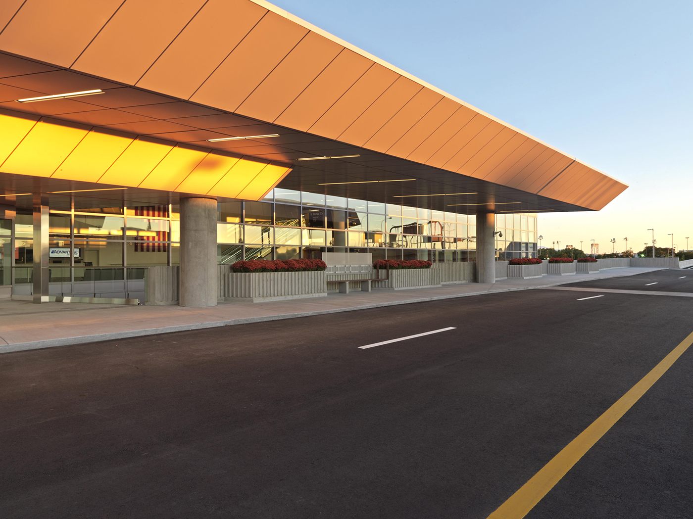 A modern airport terminal exterior at sunset, with a wide road in front, glass walls, and a large orange overhang reflecting sunlight. Red flowers line the entrance area.