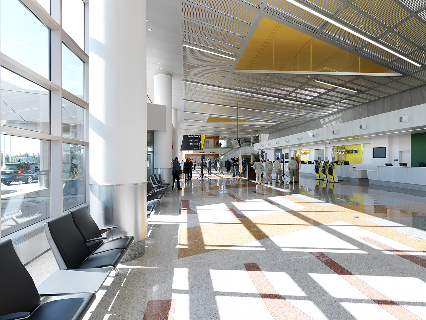 A spacious, sunlit airport terminal with rows of black seats, large windows, people walking, and check-in counters on the right side. Signs hang from the ceiling, and natural light fills the area.
