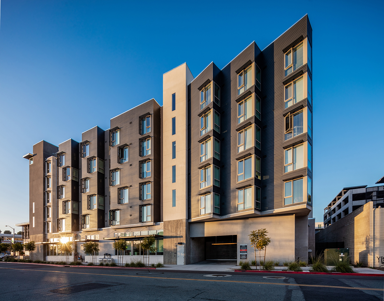 Modern five-story apartment building with sharp geometric lines, large windows, and a mix of gray exterior materials, photographed at sunset from a street corner.