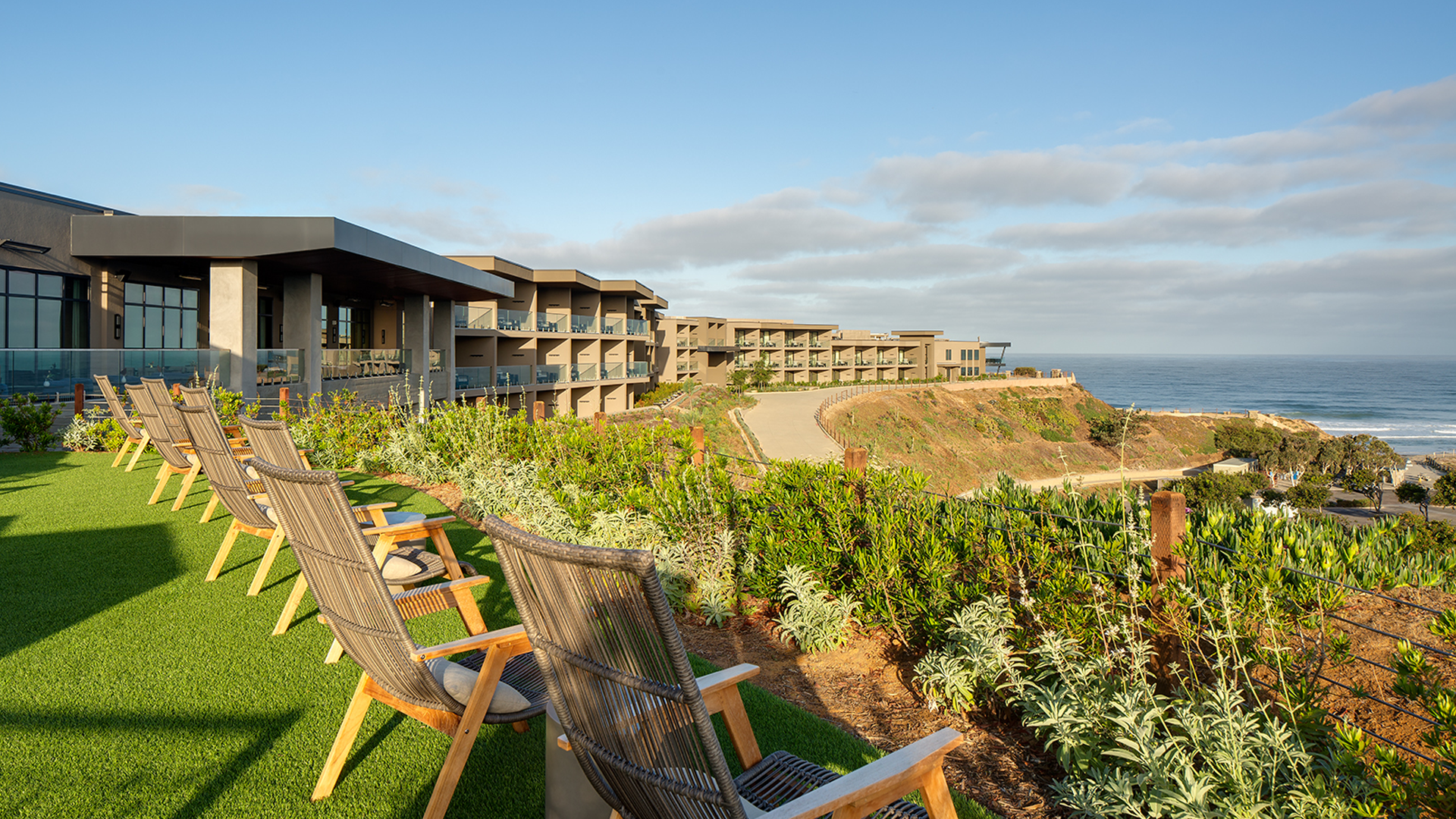 Wooden chairs line a grassy terrace overlooking a modern cliffside resort with ocean views, under a partly cloudy sky.