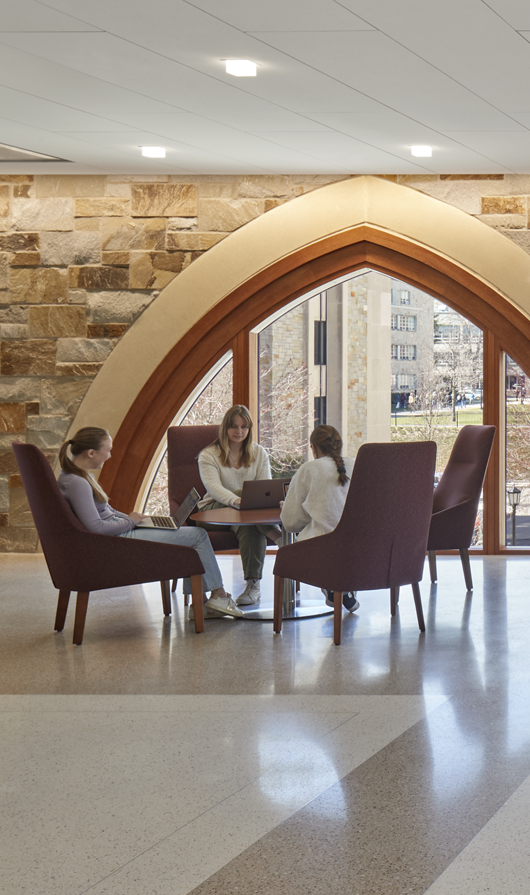A modern, sunlit study area with large windows, stone walls, and arched wooden accents. People sit at tables and workstations, while one person walks down the stairs near the windows.