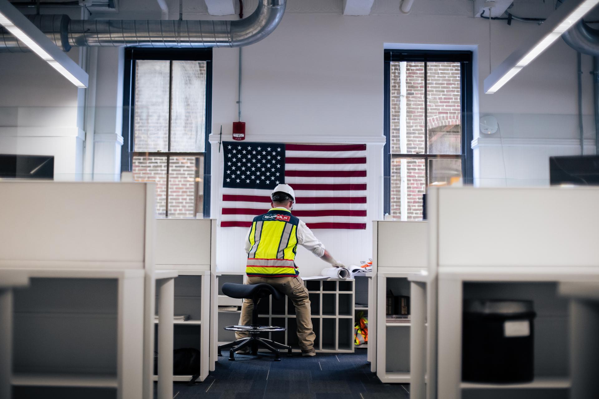 A construction worker in a safety vest and helmet sits at a desk in an office space, facing an American flag hanging on the wall between two large windows—a proud symbol for those pursuing careers in construction.