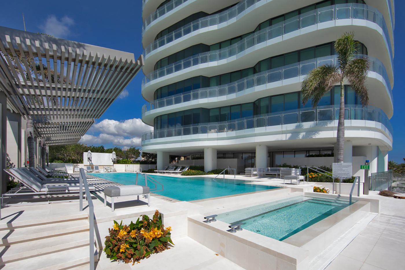 Luxury apartment building with modern curved balconies, overlooks a pristine outdoor pool area. Lounge chairs, palm tree, and lush landscaping are visible under a bright blue sky with scattered clouds.