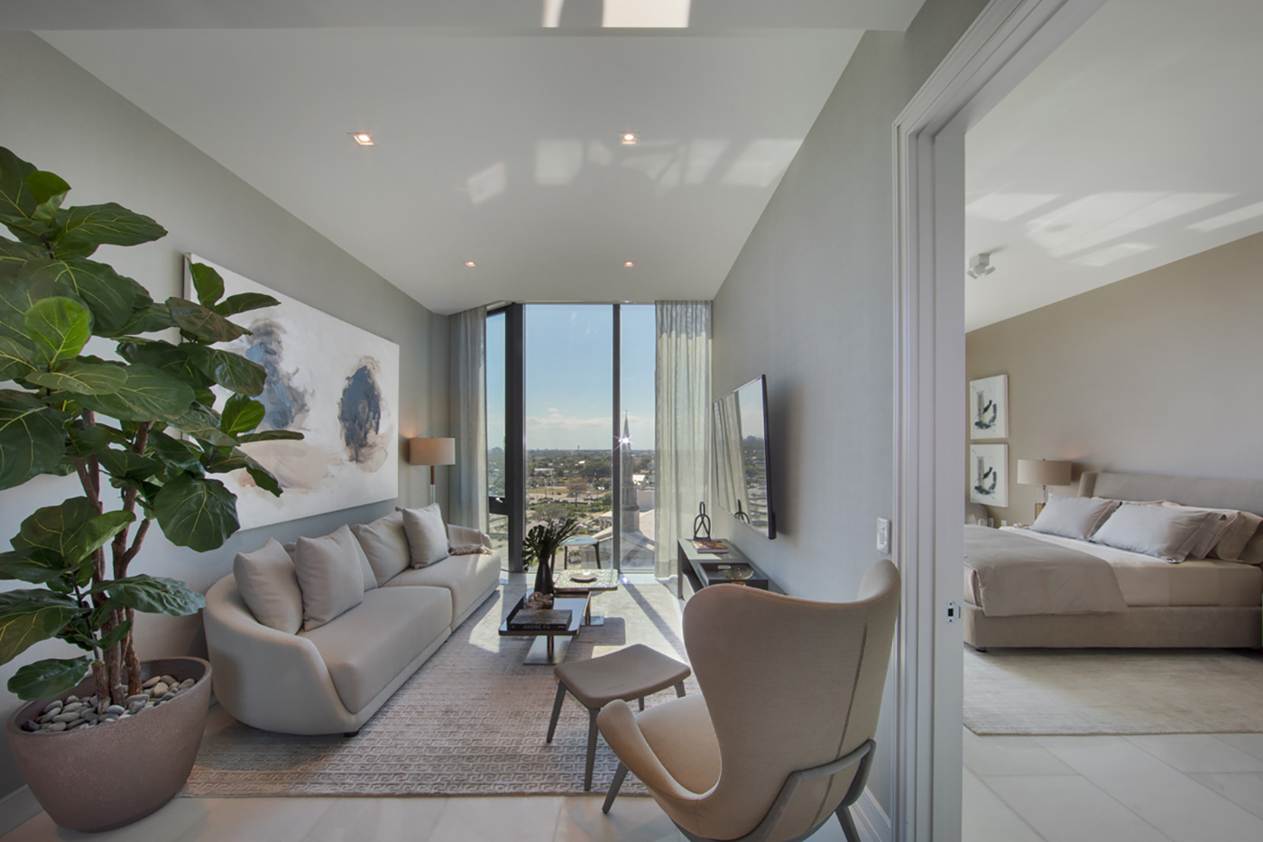 Modern living room with light-gray sofa, abstract art, large potted plant, lounge chair, and floor-to-ceiling windows overlooking a cityscape; adjacent bedroom visible through open sliding door.