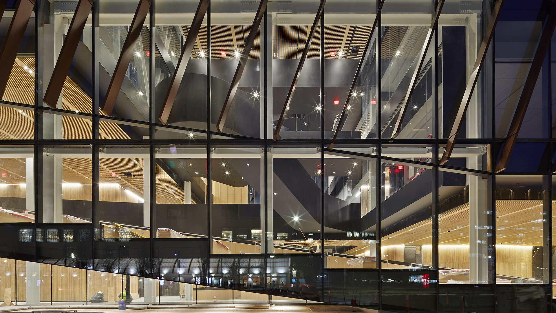 A modern glass home at night reveals brightly lit interiors, with visible black staircases, wooden ceilings, and scattered lights, reflecting the exterior city lights on the transparent facade.