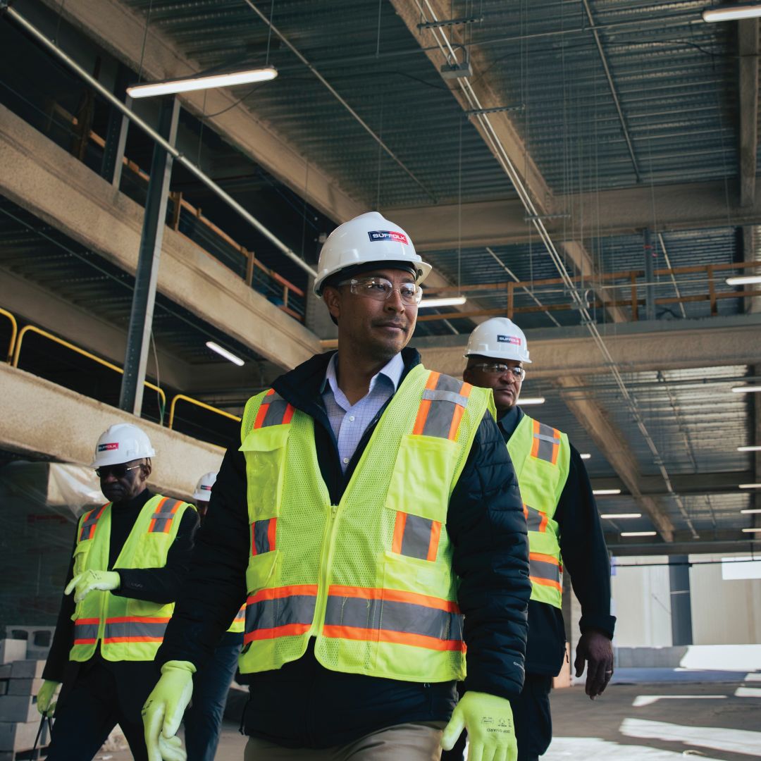 A group of construction workers wearing hard hats, safety vests, and gloves walk through a partially built commercial building with exposed steel and concrete, embodying a strong sense of community on the job site.