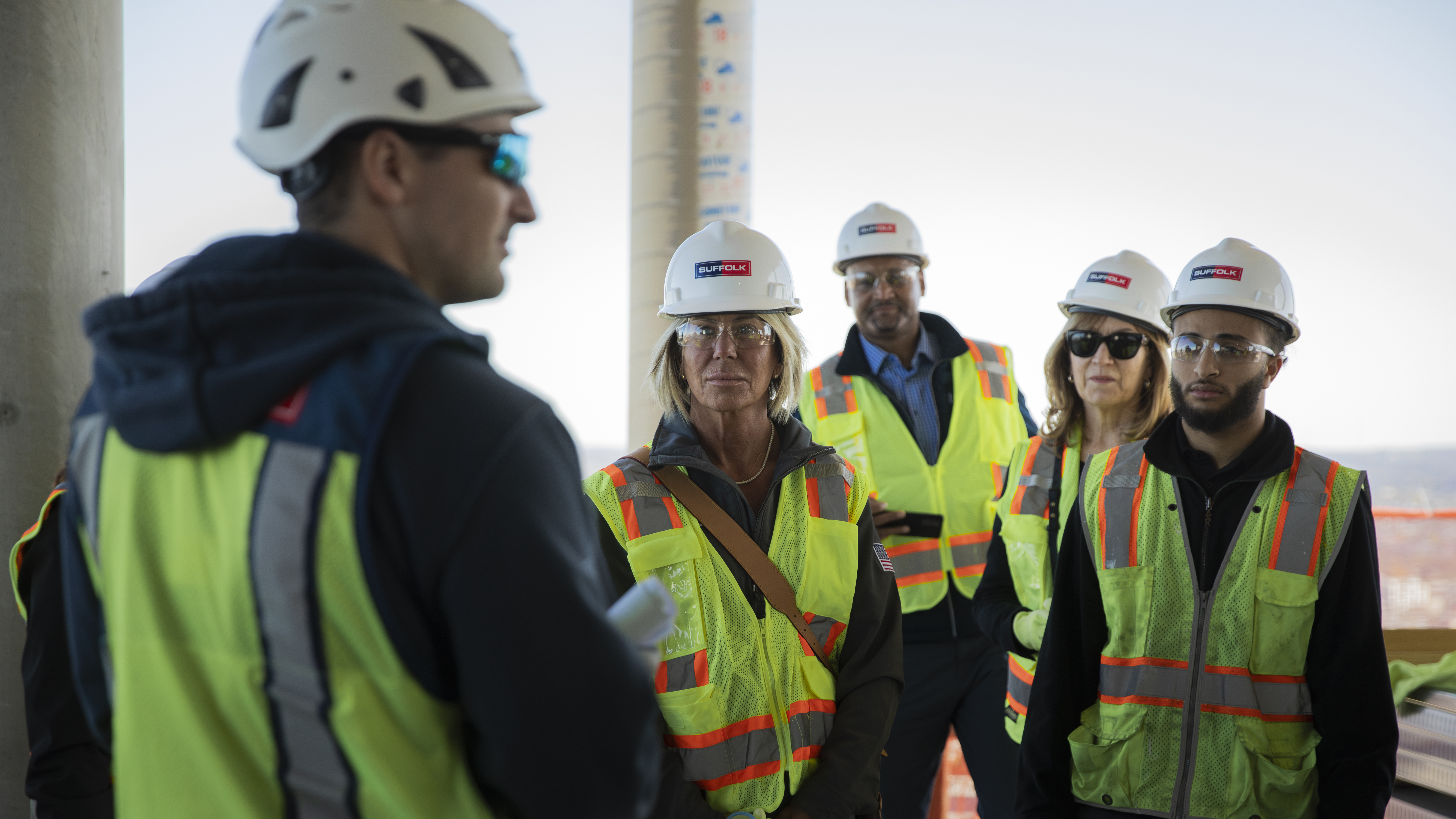 A group of five people wearing safety vests, hard hats, and glasses listen attentively to a person speaking at a construction site. The background shows the unfinished structure and bright daylight.