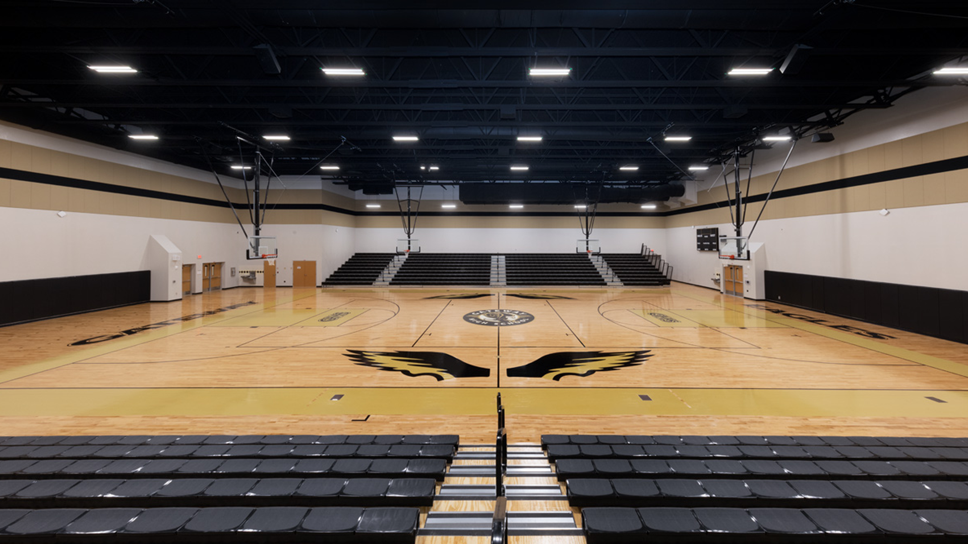 Wide view of an empty indoor basketball gymnasium with bleachers on both sides, wooden flooring, and basketball hoops at each end. A large winged emblem is at center court. The gym is well-lit with overhead lights.