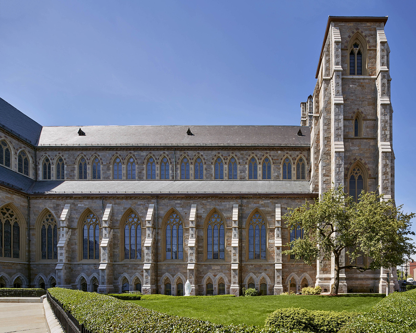 A large stone building with tall arched windows and a prominent tower, surrounded by a manicured lawn and hedges under a clear blue sky.