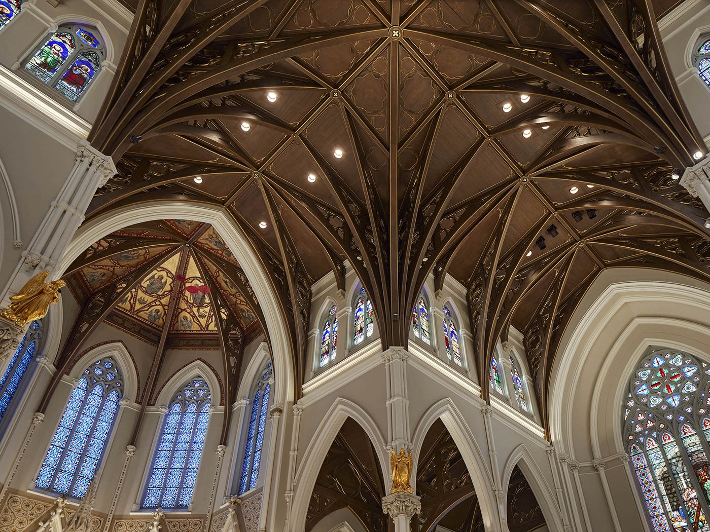 The image shows the ornate vaulted ceiling and stained glass windows of a grand Gothic-style cathedral, with pointed arches and decorative details. Warm lighting highlights the dark wood ceiling and intricate architectural features.