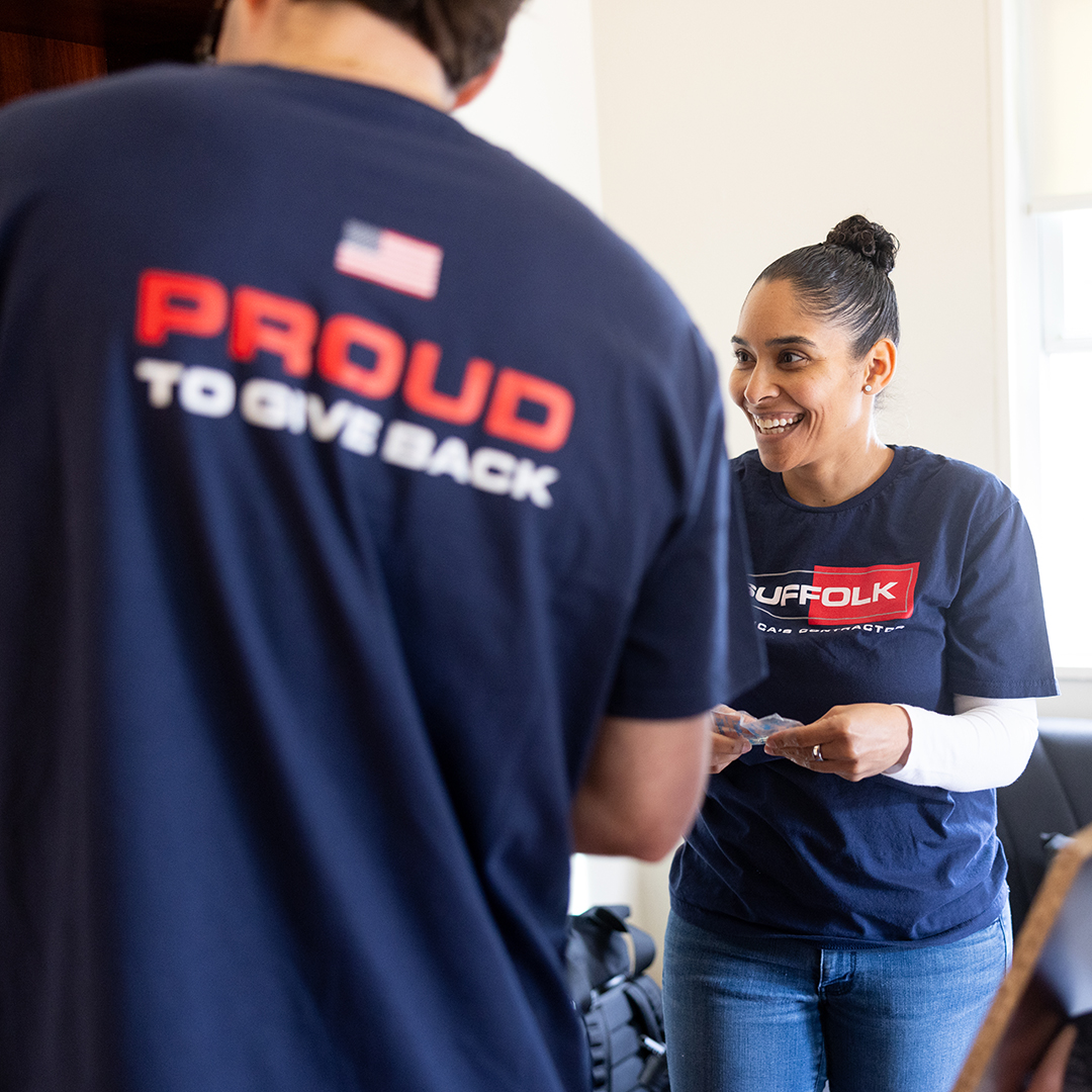 Two people wearing blue Suffolk shirts interact, embodying community spirit. The woman smiles while holding papers; the man’s back displays “Proud to give back” in bold letters as they volunteer or work together.