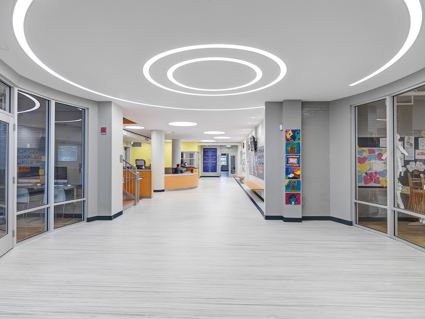 A bright, modern hallway with white floors, circular ceiling lights, and glass-walled rooms on both sides. Colorful artwork is displayed on walls, and an orange reception desk is seen in the background.