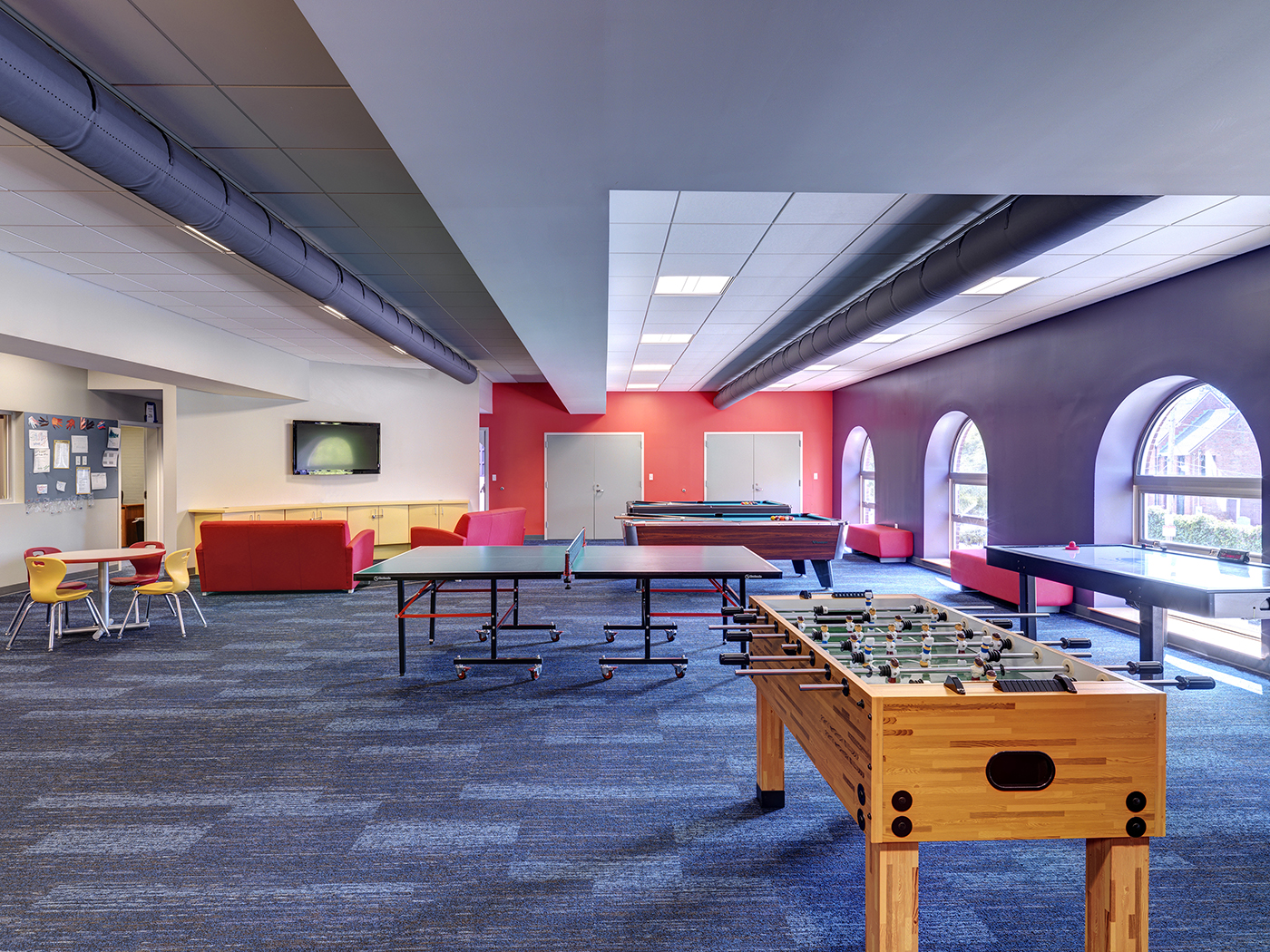 A modern rec room with blue carpet, red and white walls, ping pong and foosball tables, lounge chairs, TV, and large arched windows letting in natural light.