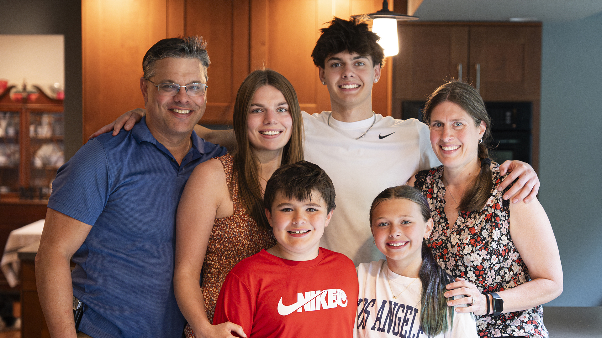 A smiling family of six poses together in a kitchen, reflecting the warmth and togetherness that defines their culture. Two adults stand on each end, with four children—two boys and two girls—in between. They are all casually dressed and appear happy.