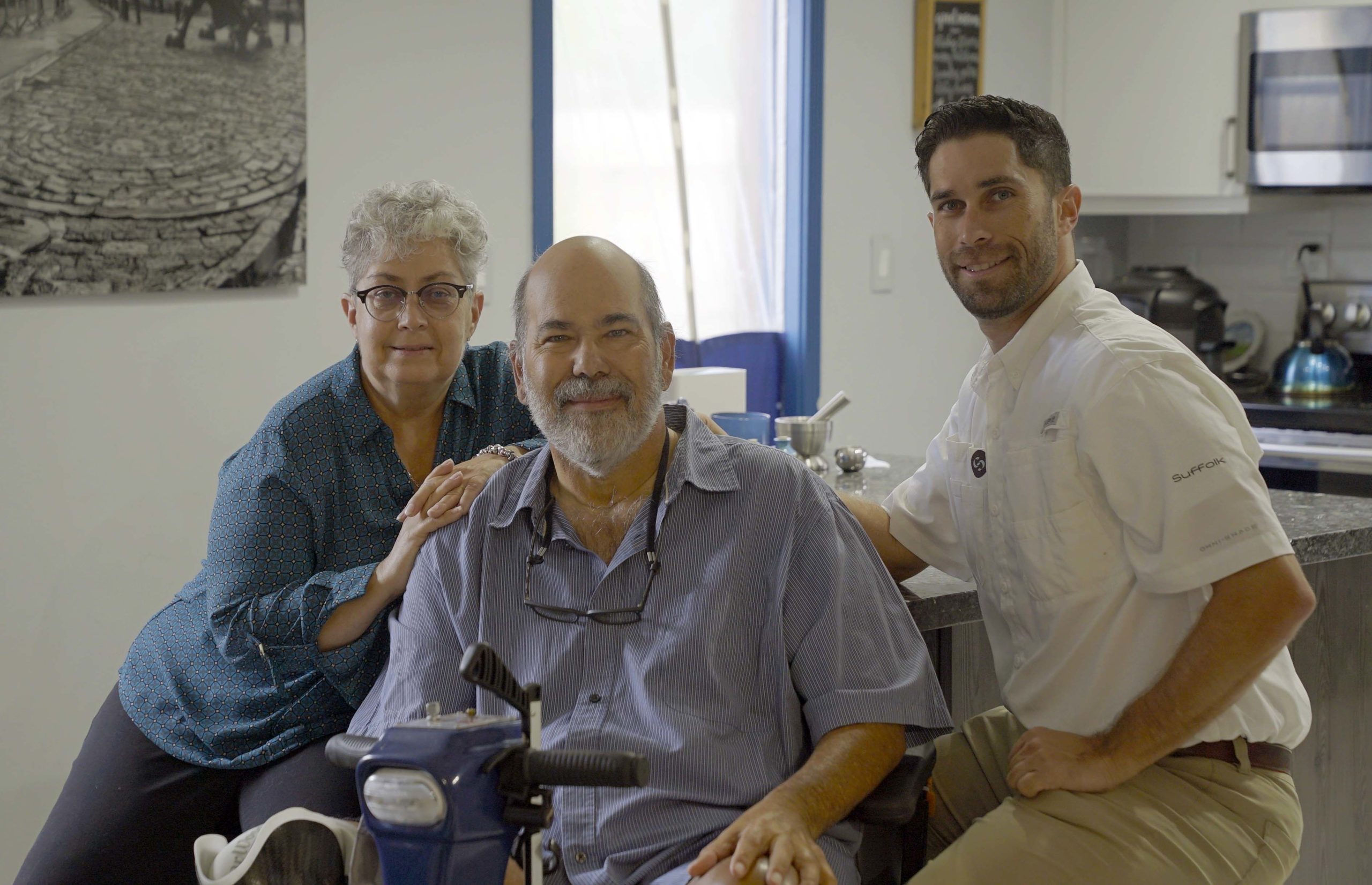 Three people smile together in a kitchen: an older woman with gray hair and glasses stands beside an older man sitting on a mobility scooter, while a younger man kneels next to them. They appear happy and relaxed.