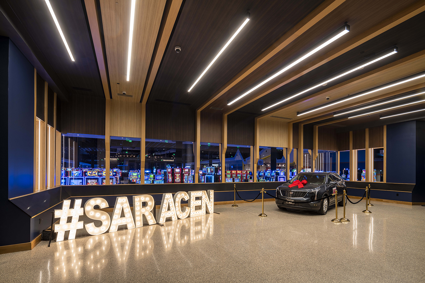 A sleek black car with a large red bow is displayed behind ropes next to a lit-up #SARACEN sign; slot machines and bright lights are visible in the background, creating a casino atmosphere.