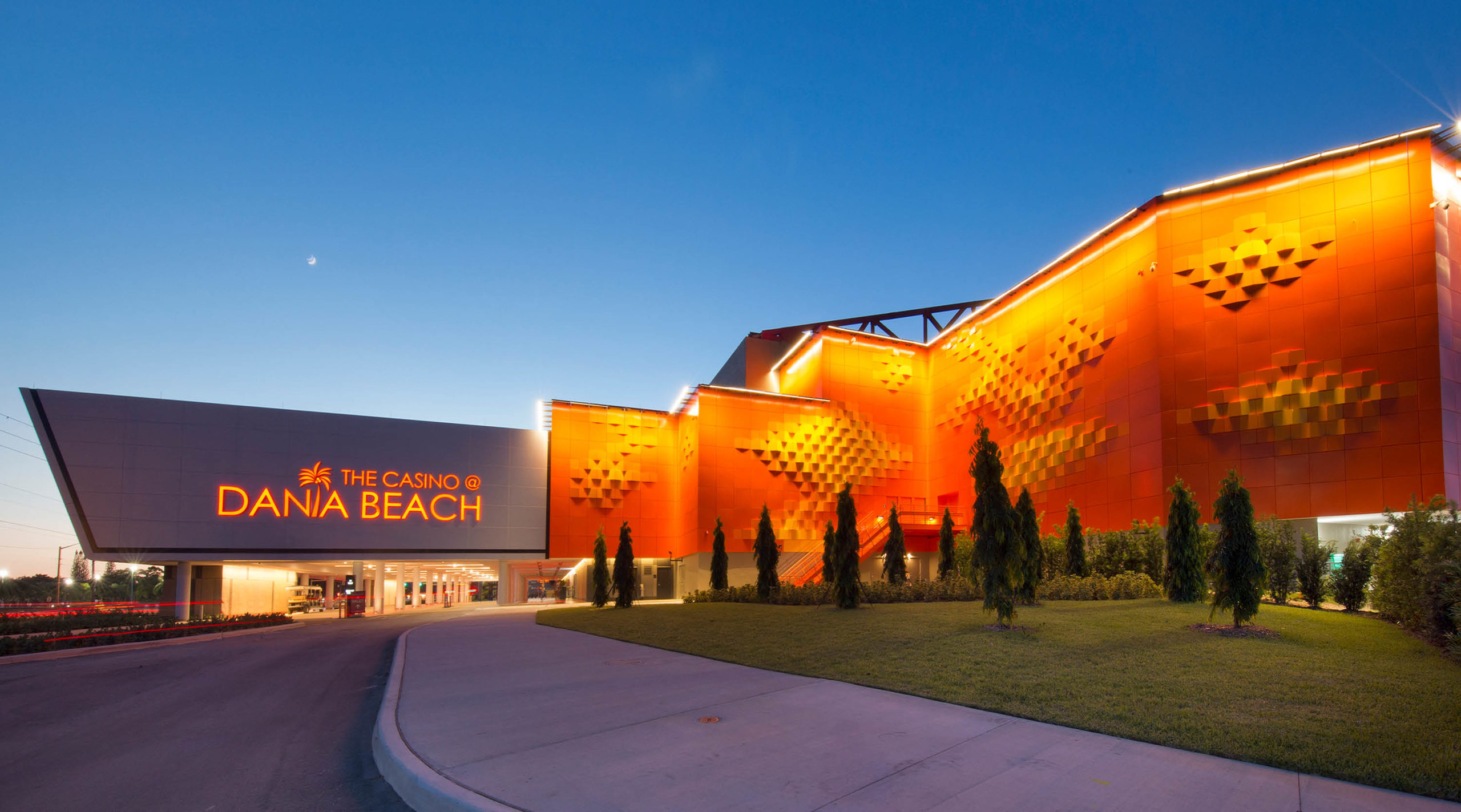 Bright orange and modern exterior of The Casino at Dania Beach at dusk, with illuminated signage, geometric wall patterns, a manicured lawn, and a clear evening sky above.