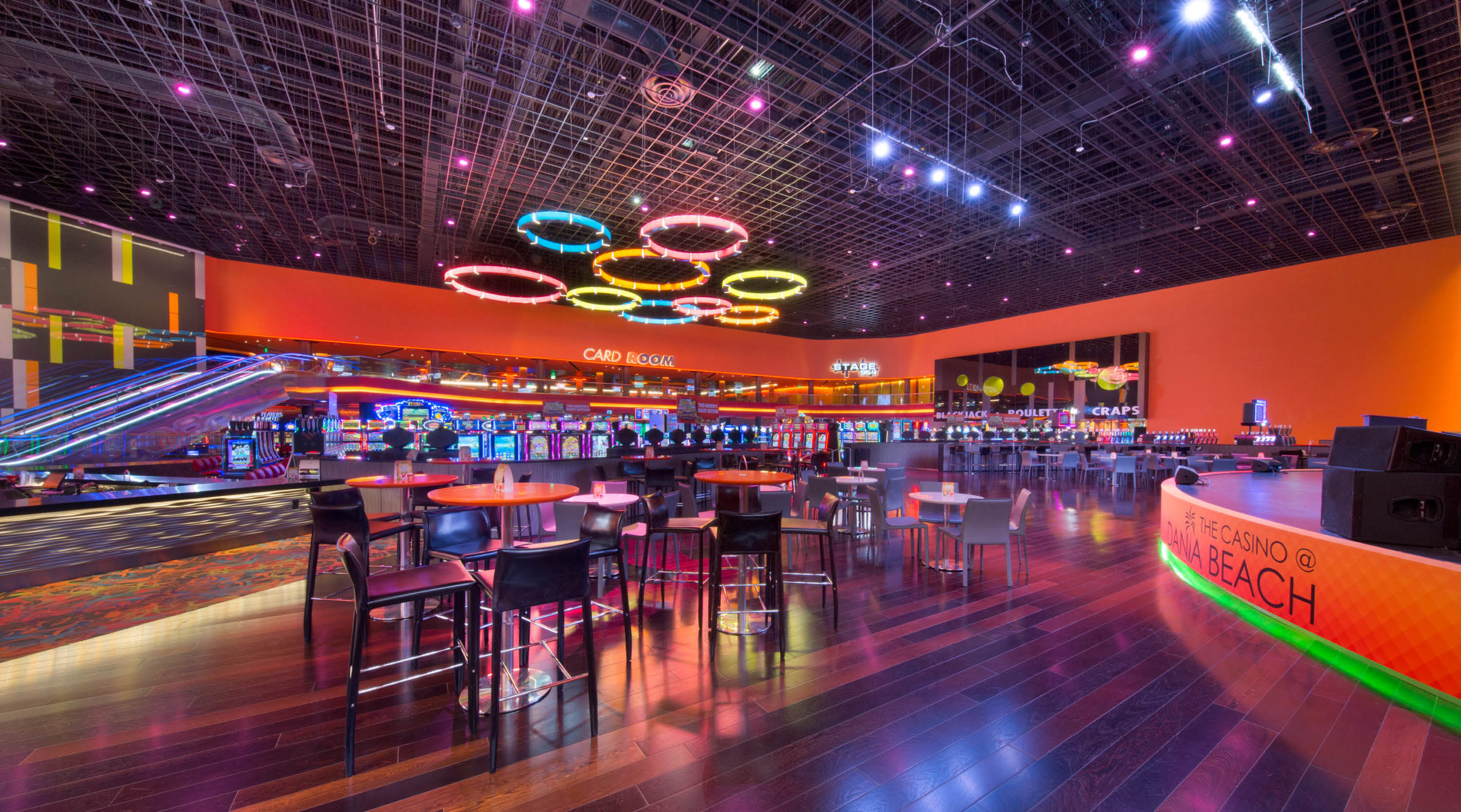 A brightly lit, modern casino interior with colorful neon ceiling lights, slot machines, high-top tables and chairs, and an orange and yellow color scheme. The space is spacious and vibrant.