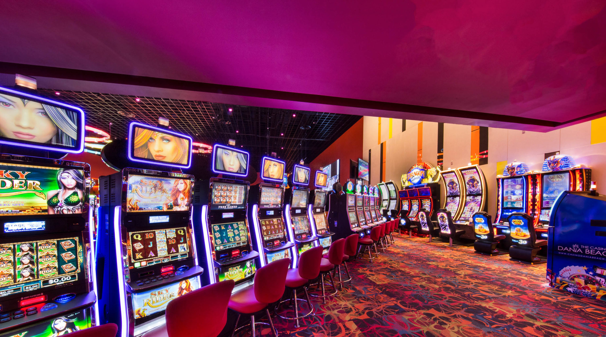 A brightly lit casino floor with rows of colorful slot machines and red chairs on patterned carpet under a glowing magenta ceiling. Screens on the machines display various games and animated graphics.