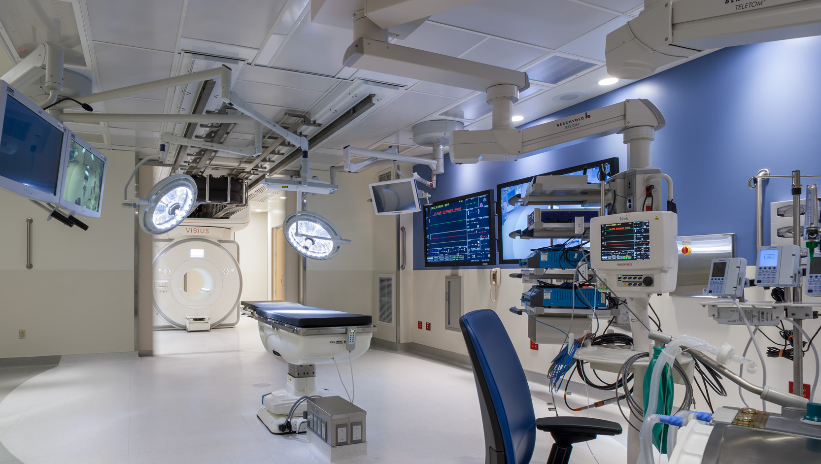 A modern hospital operating room features advanced medical equipment, large monitors displaying vital signs, a surgical table, and bright overhead lights. There is also an MRI machine visible in the background.