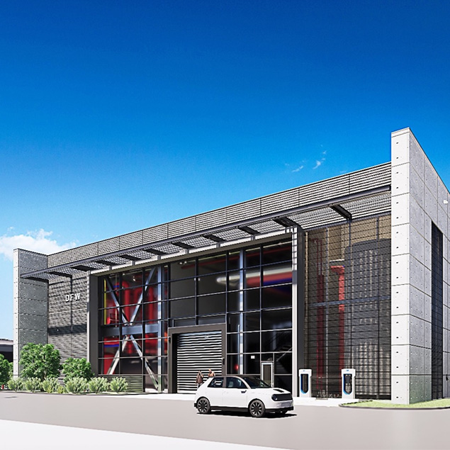 Modern industrial building with a glass facade and metal accents, featuring large windows, a parked white car in front, electric vehicle charging stations, and clear blue sky overhead.