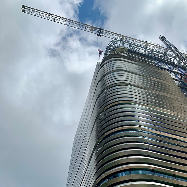 A modern high-rise building under construction with a large crane on top, set against a cloudy sky. The building features curved, metallic lines and reflective surfaces.
