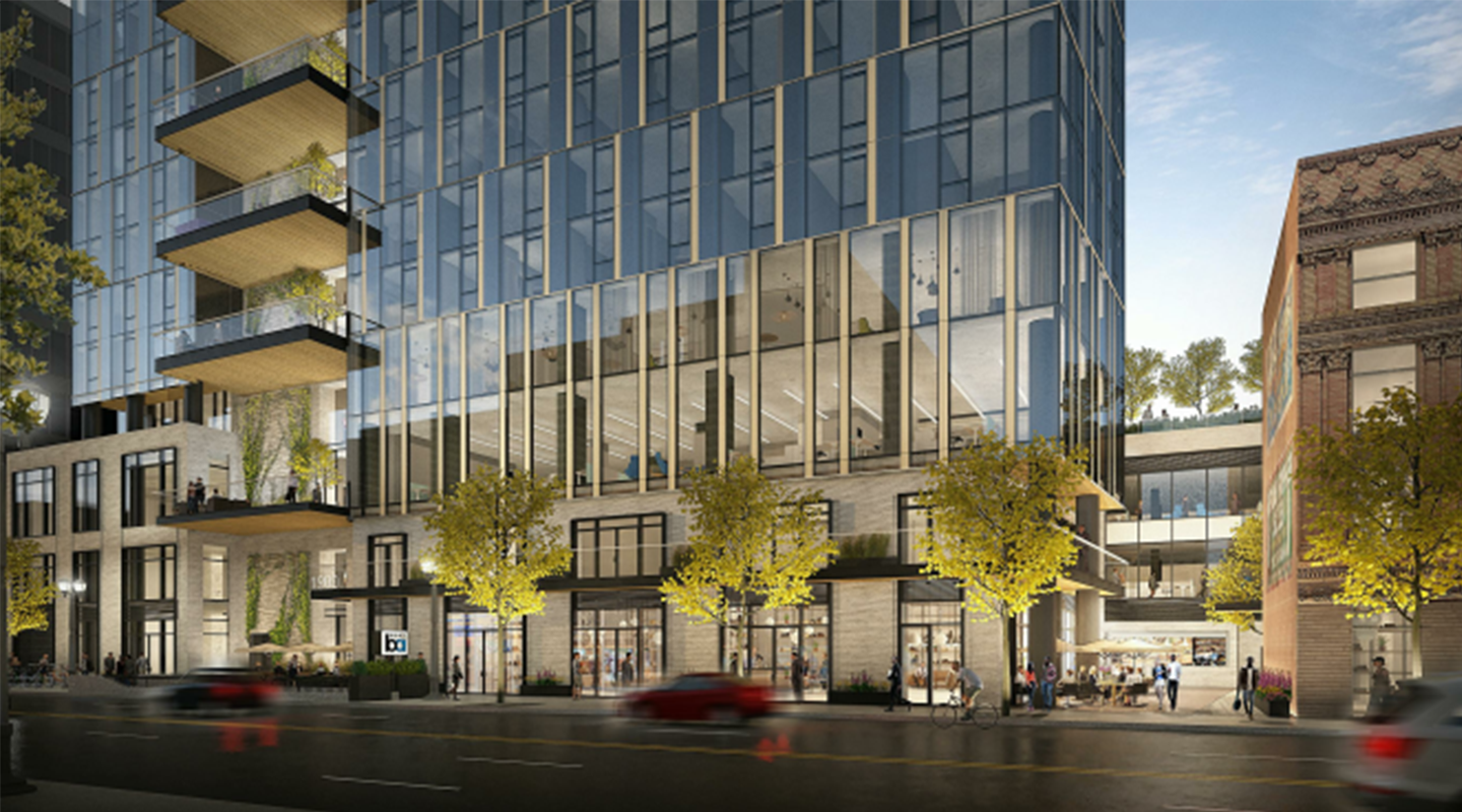 Modern glass office building with large windows and balconies, surrounded by trees and street-level shops. Cars and pedestrians are visible on the busy street in front. Adjacent brick building is partially visible on the right.
