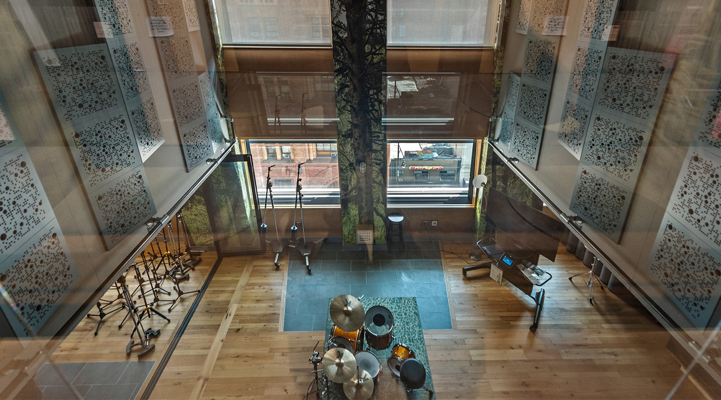 A modern music recording studio viewed from above, featuring a drum set, microphones, a piano, wooden floors, large windows, and decorative sound panels on the walls.